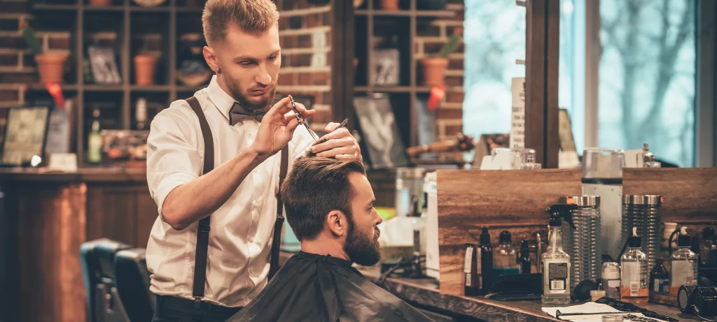 Young bearded man getting haircut by hairdresser while sitting in chair at barbershop