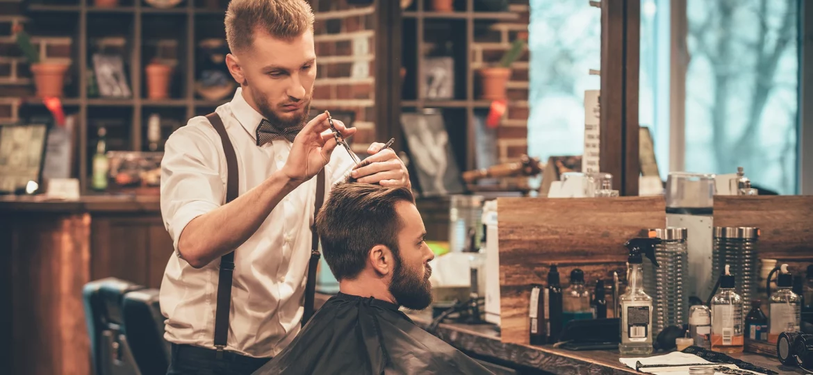 Young bearded man getting haircut by hairdresser while sitting in chair at barbershop