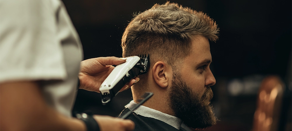 Young bearded man getting haircut by hairdresser while sitting in chair at barbershop